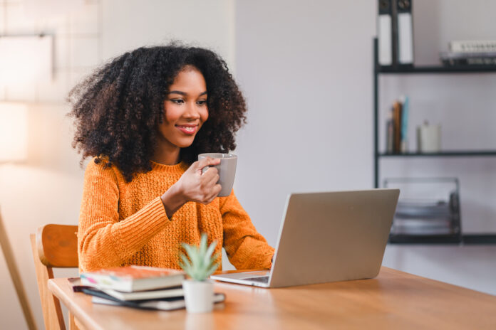 Focused African American woman holding a coffee mug while working on a laptop at home, sitting at a desk with books.